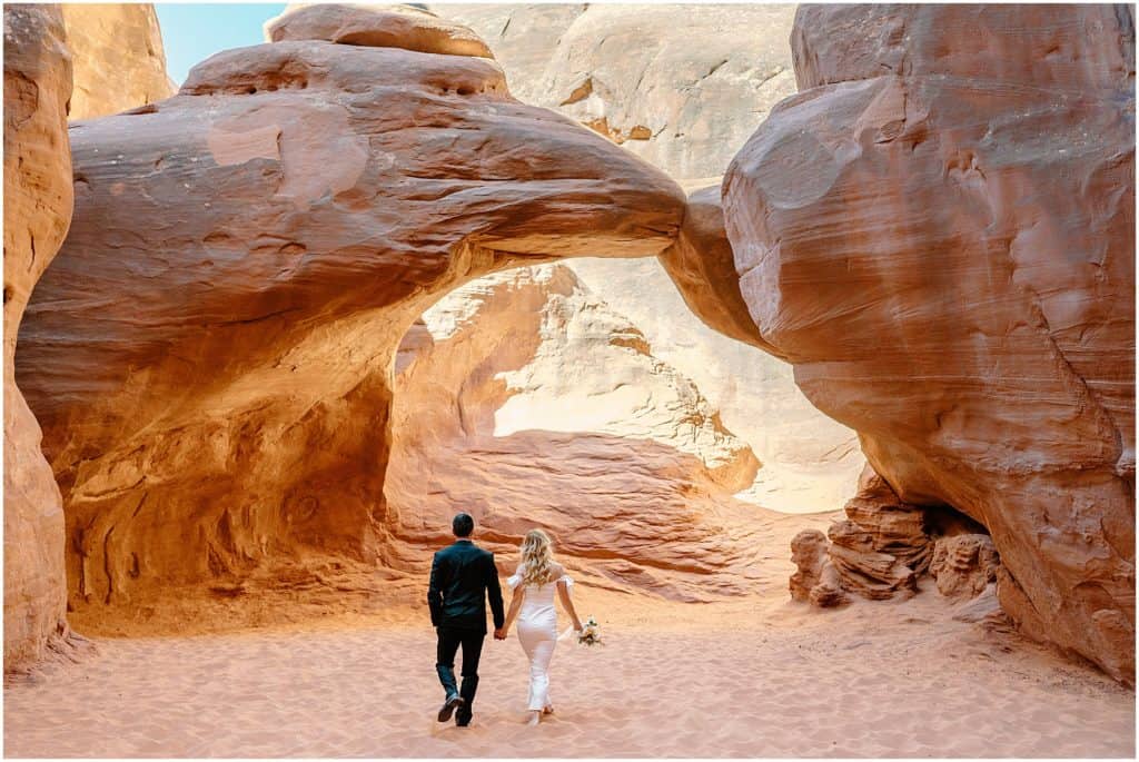 couple walking in sand. under an arch on their moab elopement day.