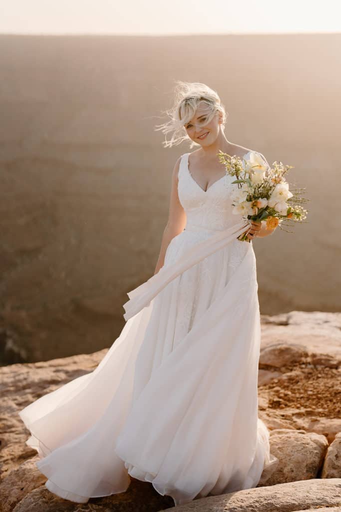 A bride in a white elopement dress lets the light fabric blow in the breeze on an Utah cliffside. 