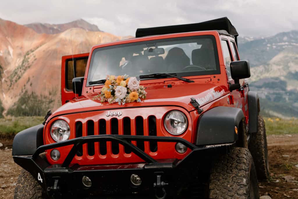 An orange jeep is shown against a dramatic, mountainous backdrop. A couple, Ana and Greg, rented this jeep for their Colorado elopement wedding. 