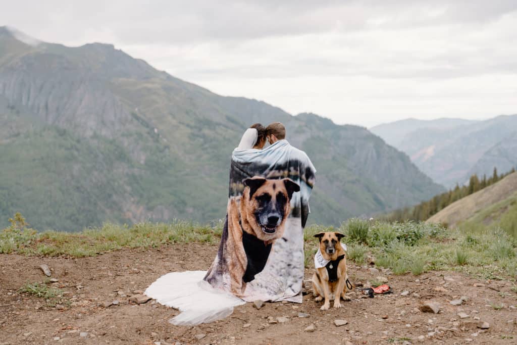 A couple wraps themselves in a blanket with an image of their German shepherd as they finish their vows during their Colorado elopement. The dog sits by them as if posing for the photo. 