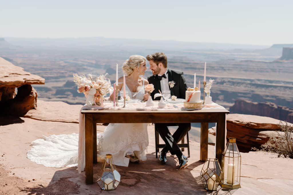 A bride and groom hold each other close as they sit behind a beautiful sweetheart table on the cliff of Dead Horse State Park during their adventure elopement ceremony.