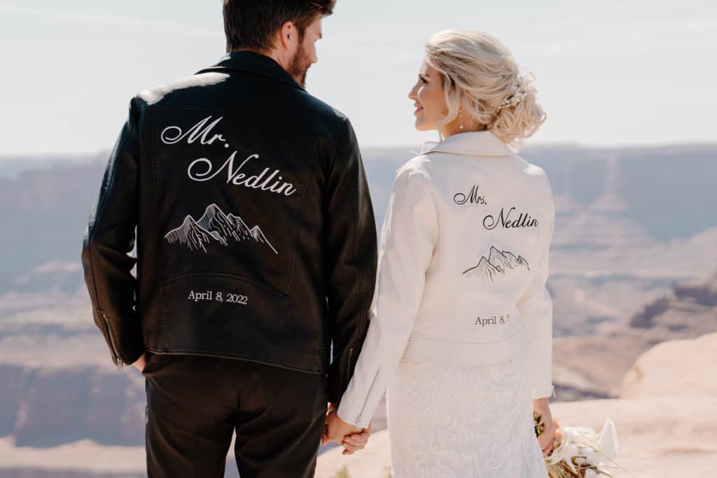 A bride and groom look out over a vista holding hands in matching leather jackets.