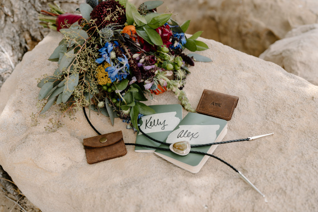 A bride and grooms wedding day details, like their vow books, boquets, and bolo tie, lay against a sandstone rock during their Green River rafting elopement ceremony. 