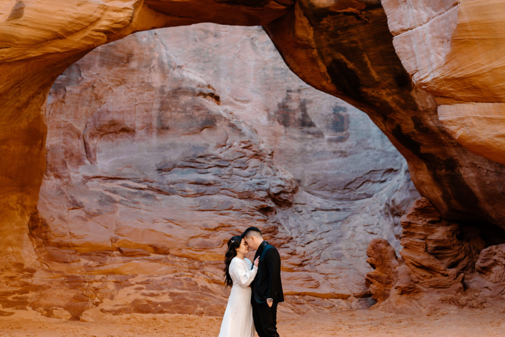 A bride and groom hold each other close underneath a delicate Utah arch on their Moab adventure elopement.