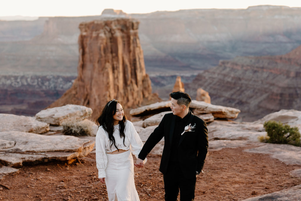 A bride and groom, in full wedding attire, hike along the rim of a gorgeous overlook during their Moab adventure elopement ceremony. 