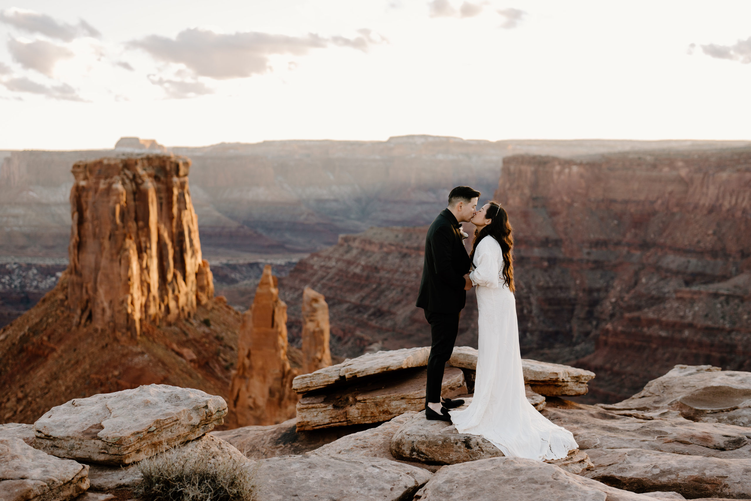 A bride and groom lean in close for a kiss during their Moab adventure elopement atop a stunning canyon overlook.