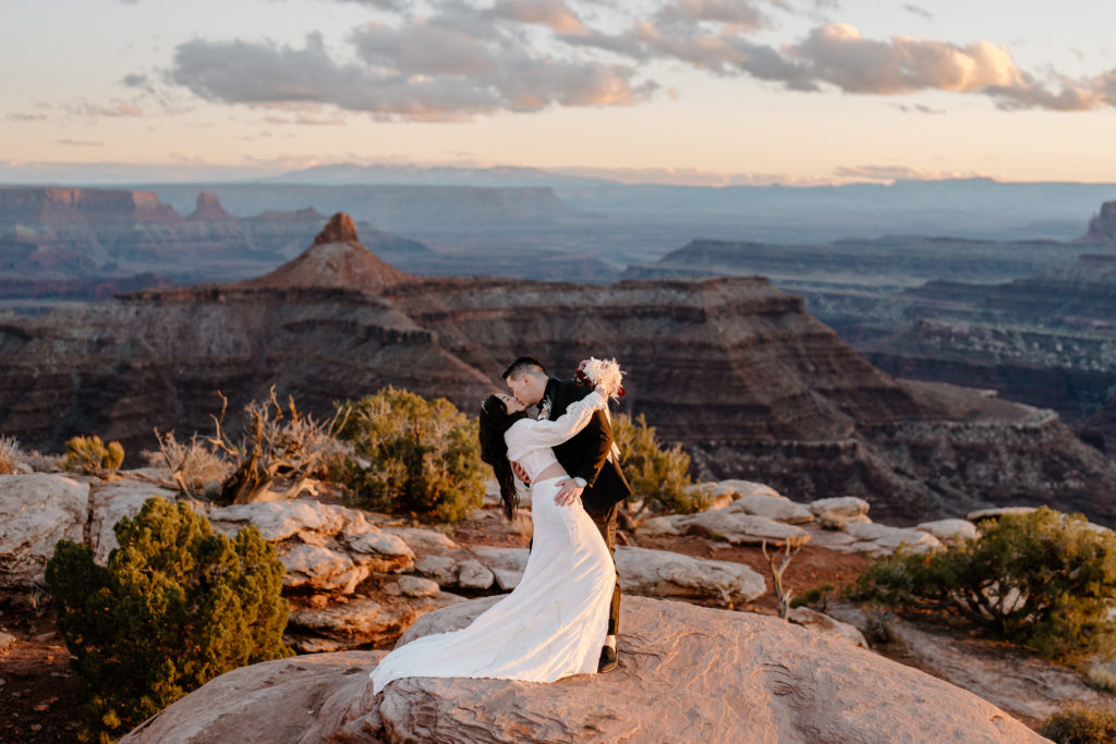 A couple, in wedding attire, kiss atop an overlook in the desert as the sun is setting during their Moab adventure elopement.