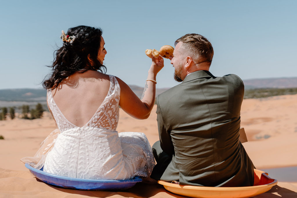 A bride and groom eat delicious donuts on sand sleds as they experience every moment of their Sand Dunes elopement.  