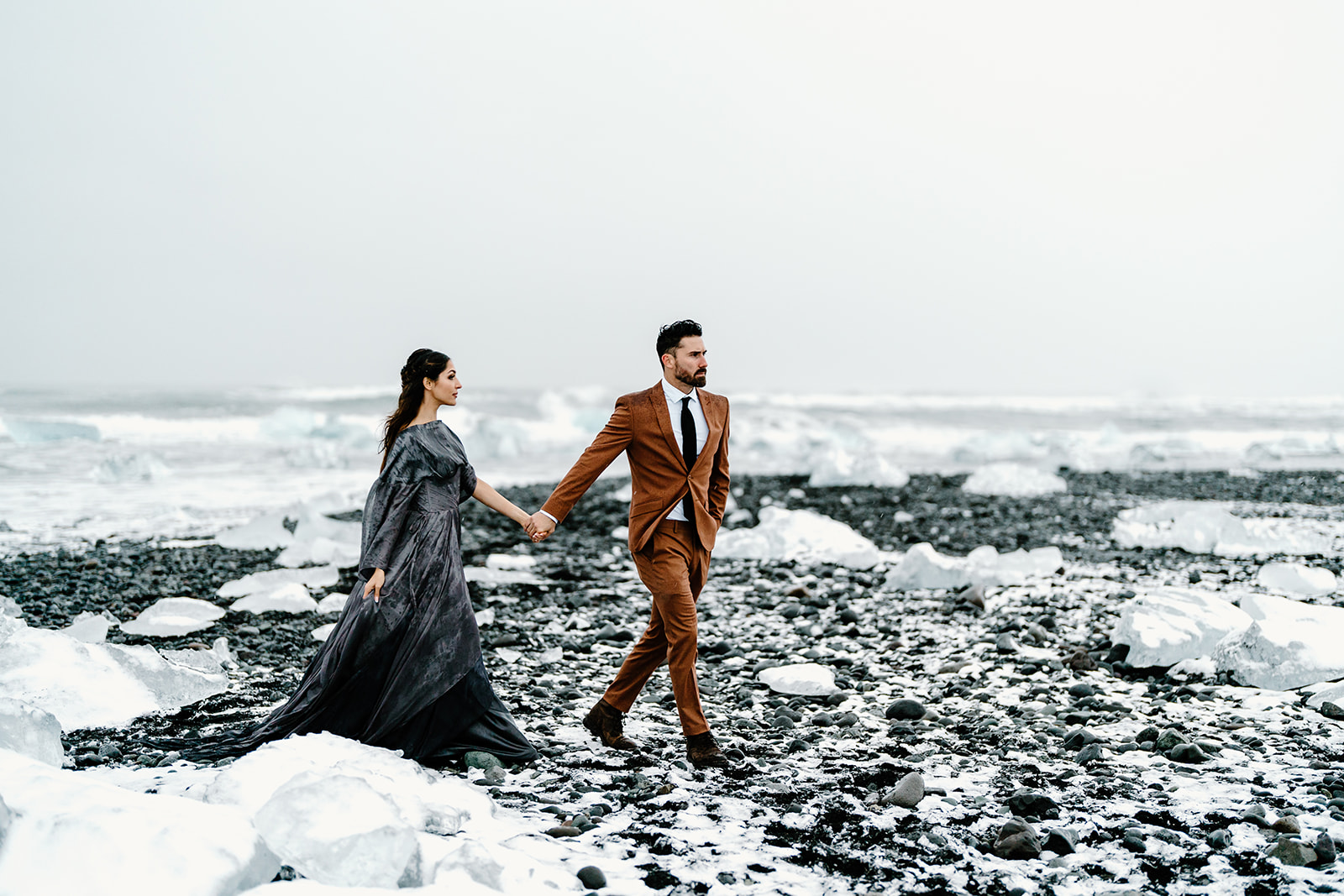 An eloping couple walks through an Icelandic ice cave in their wedding clothes.